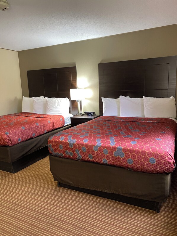 A hotel room with two beds, each with a red patterned bedspread and white pillows. There is a nightstand with a lamp and a digital clock between the beds. The room has a carpeted floor and a plain wall with a ceiling light. - SureStay by Best Western Massena (Massena)