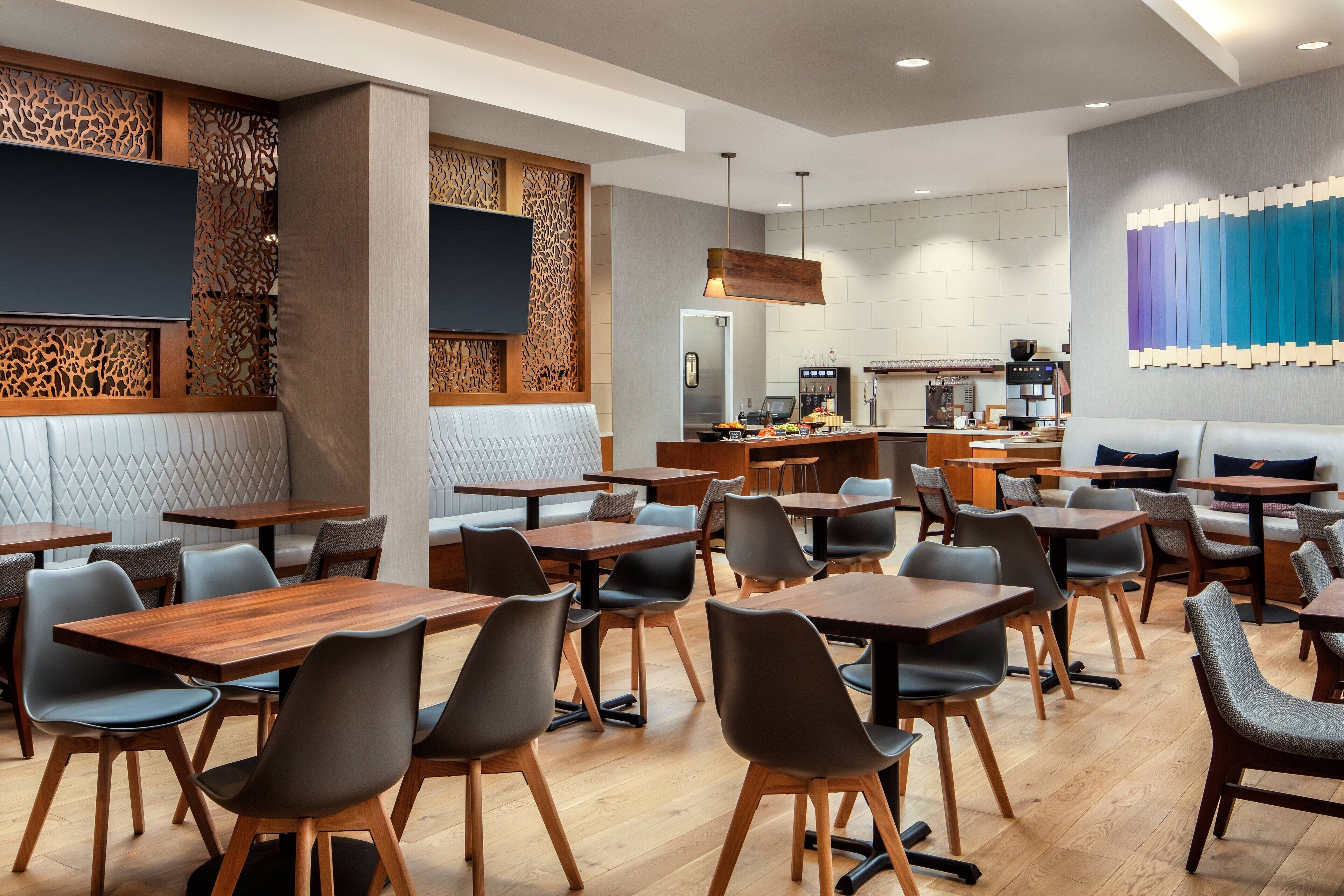 A modern restaurant with wooden tables and chairs, a kitchen in the background, and a decorative wall with a blue panel.