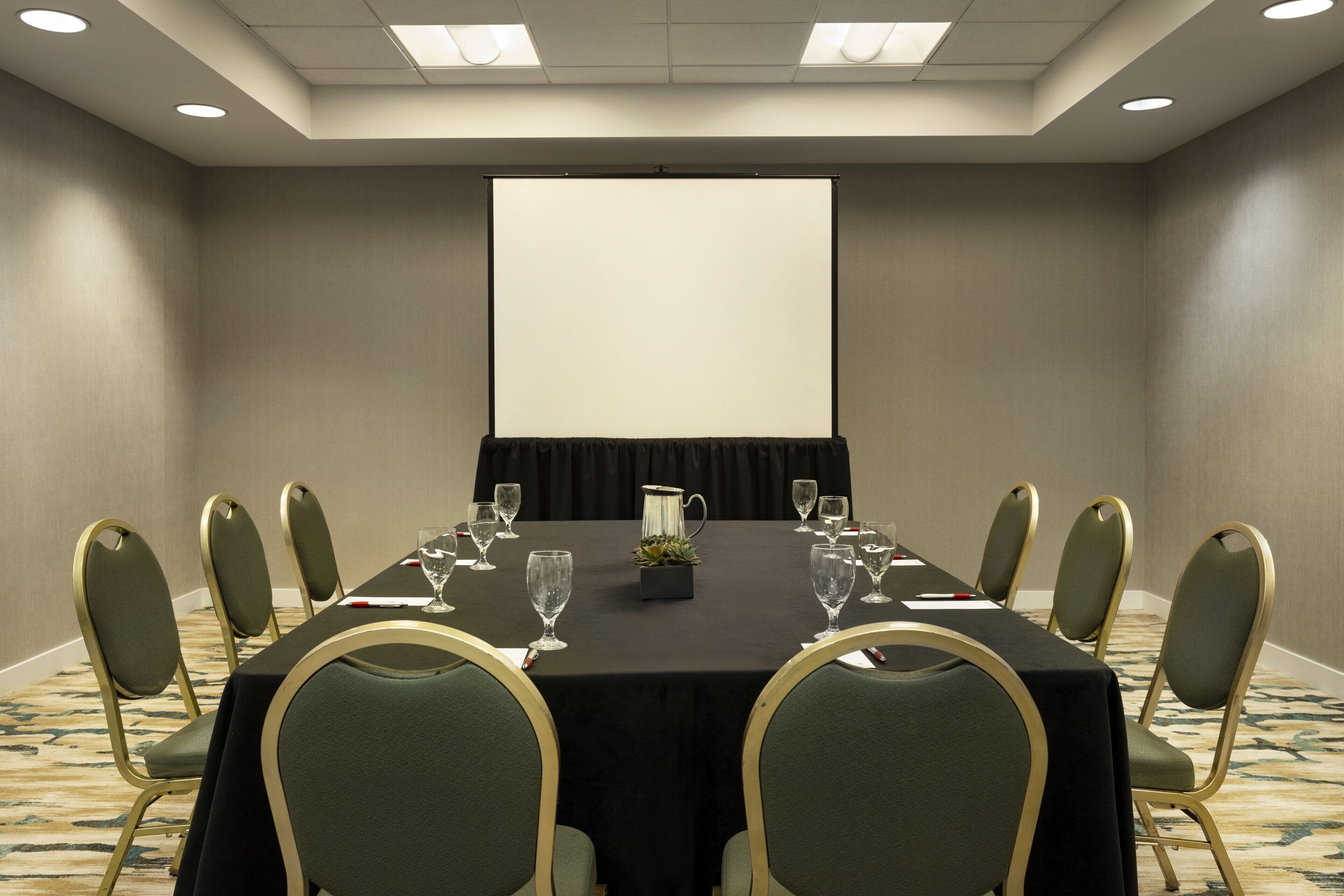 A conference room with a long table, chairs, a projector screen, and a carpeted floor.