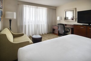 Habitación de hotel con una cama grande, un escritorio con silla, televisión y ventana con cortinas translúcidas.