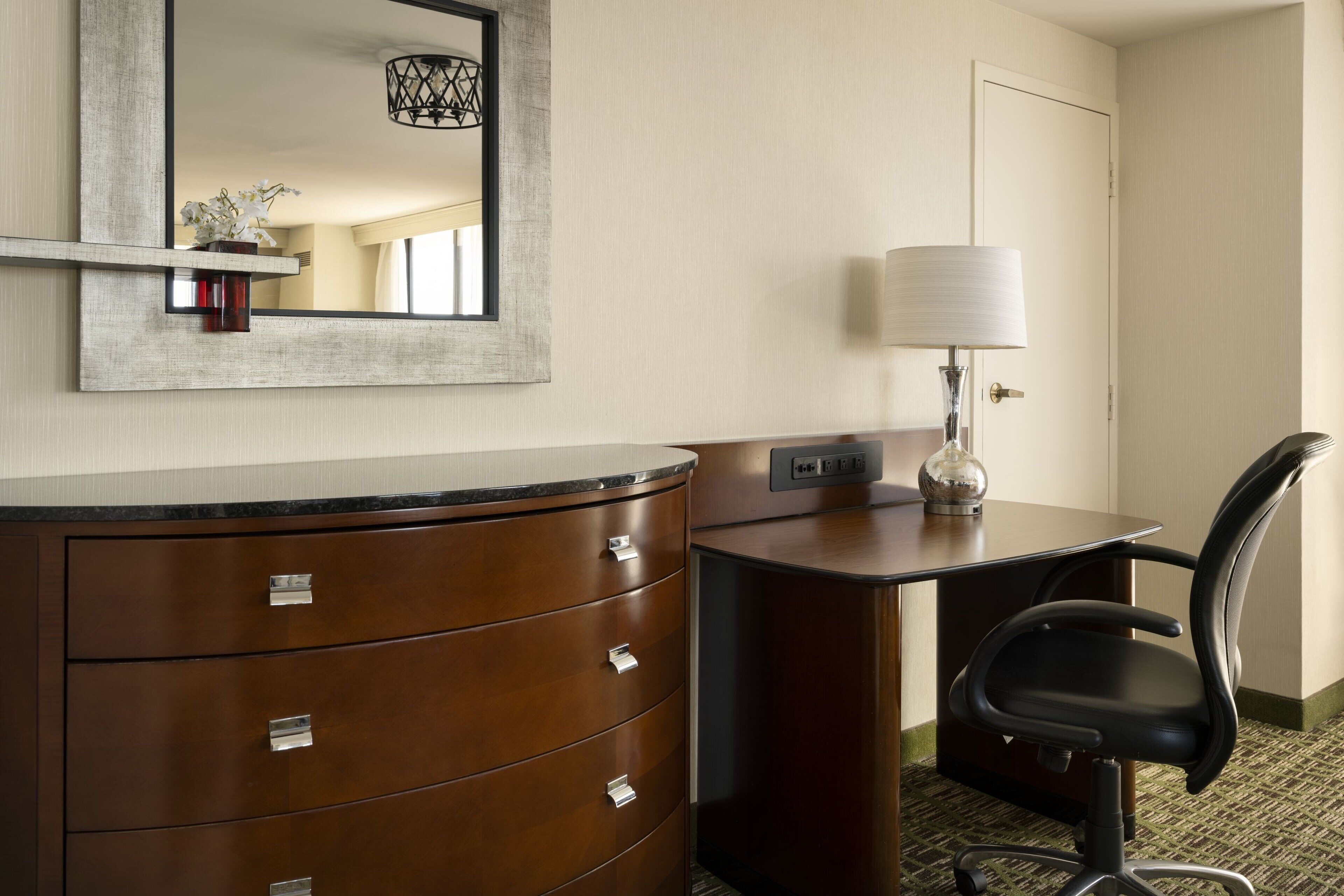 A hotel room with a wooden dresser, a desk with a lamp, a black office chair, and a mirror on the wall.