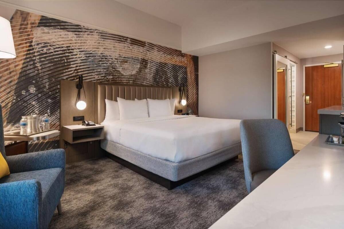 A modern hotel room with a large bed, a desk, a chair, a nightstand, and a lamp.