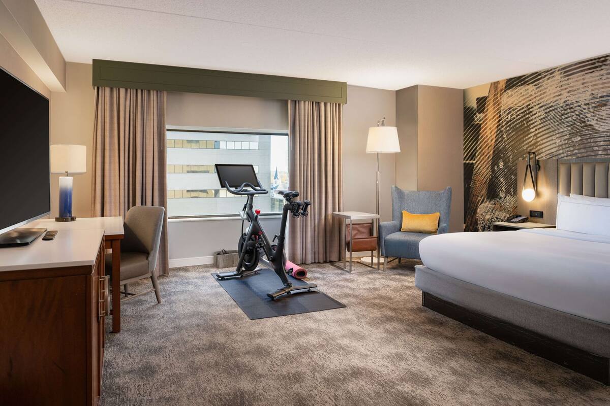 Quarto de hotel com cama, escrivaninha, cadeira, bicicleta ergométrica e vista para a cidade.