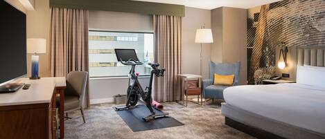 A hotel room with a bed, desk, chair, exercise bike, and a view of the city.