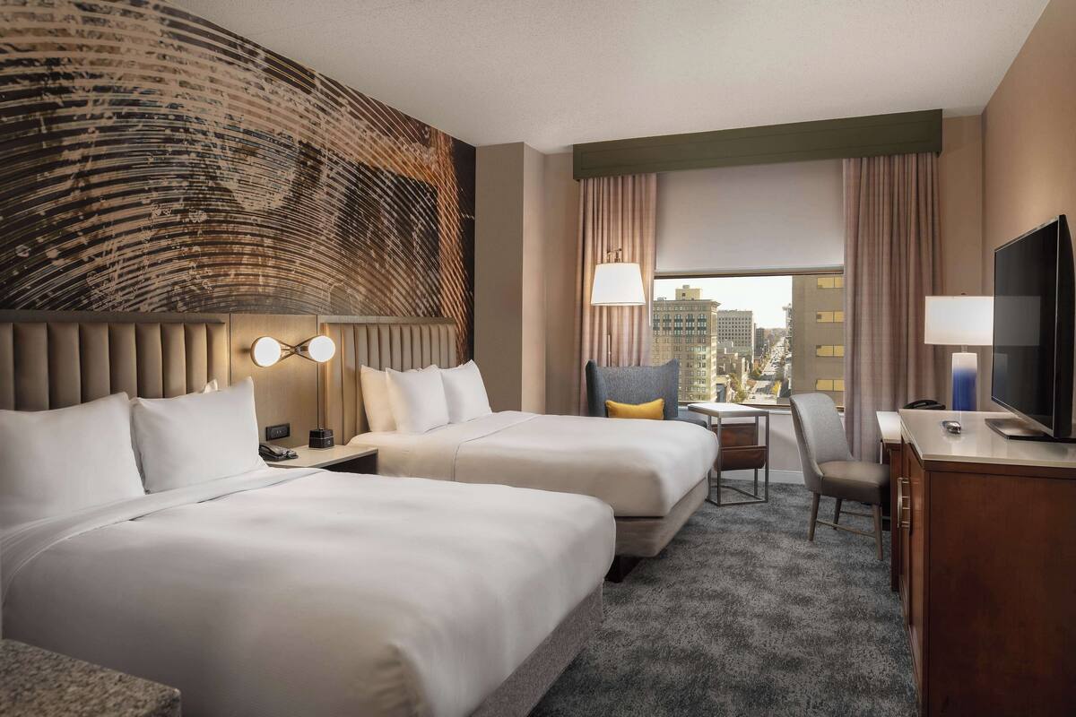 Quarto de hotel com duas camas, uma escrivaninha, uma cadeira, televisão e vista para a cidade.