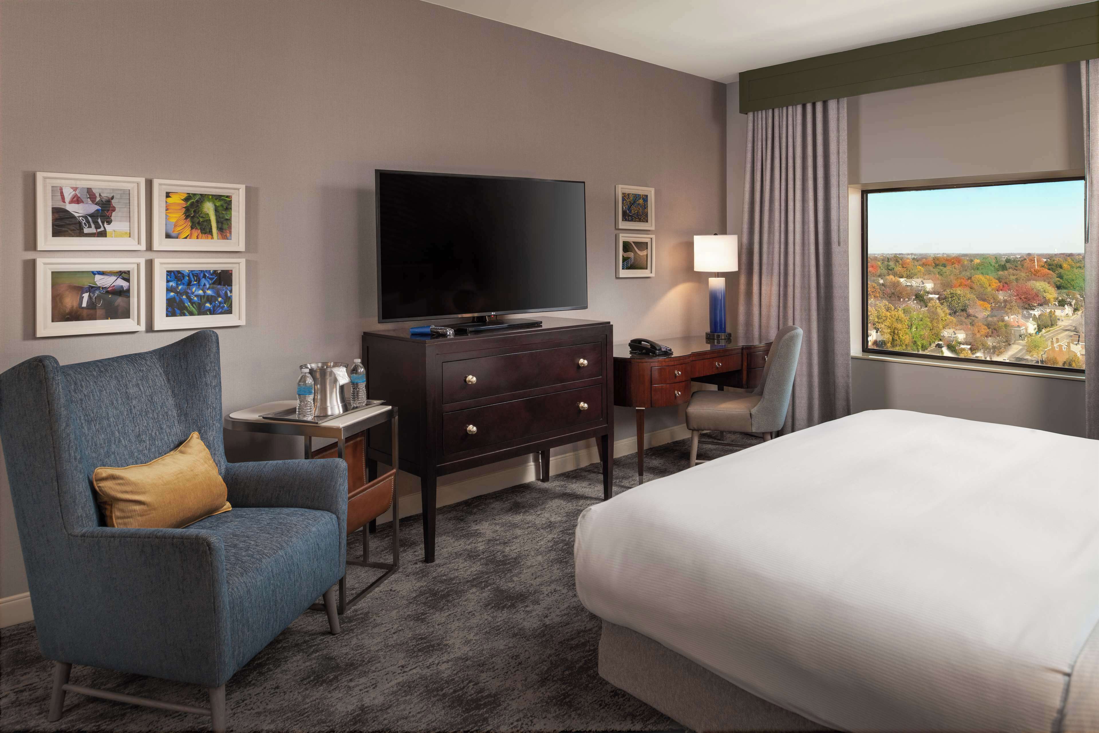 Una habitación de hotel con una cama, televisión, escritorio con lámpara, silla, cómoda y cuadros en la pared.