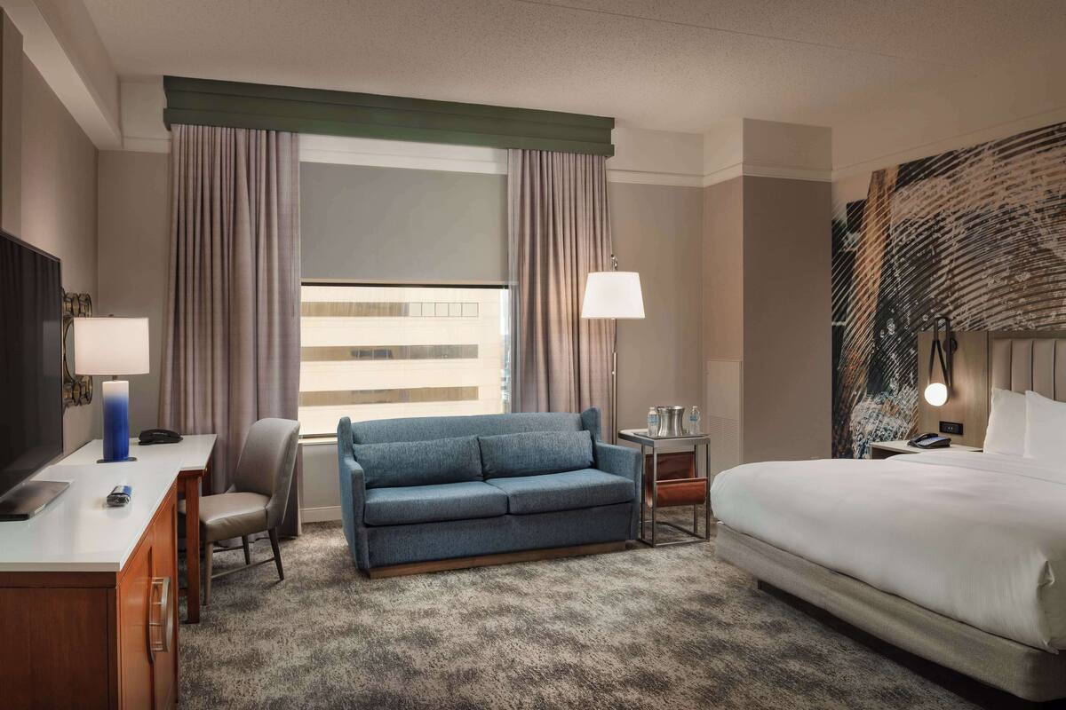 Quarto de hotel com uma cama, um sofá, uma escrivaninha com uma luminária, uma televisão e uma janela com cortinas.