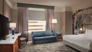 Quarto de hotel com uma cama, um sofá, uma escrivaninha com uma luminária, uma televisão e uma janela com cortinas.