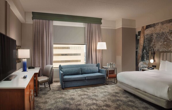 A hotel room with a bed, a sofa, a desk with a lamp, a TV, and a window with curtains.