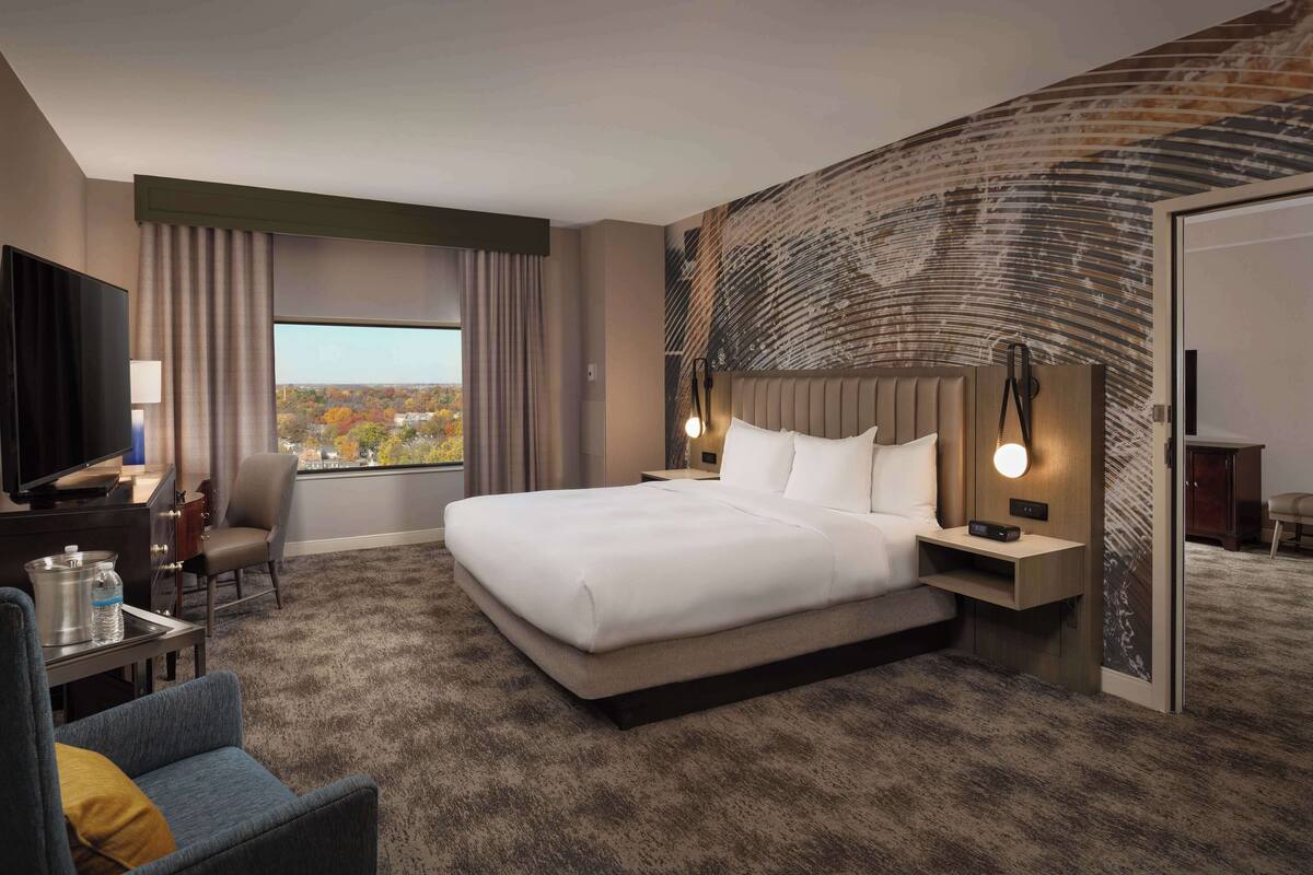 Quarto de hotel com uma cama grande, televisão, secretária com cadeira, vista para a cidade e parede de destaque em tijolo.