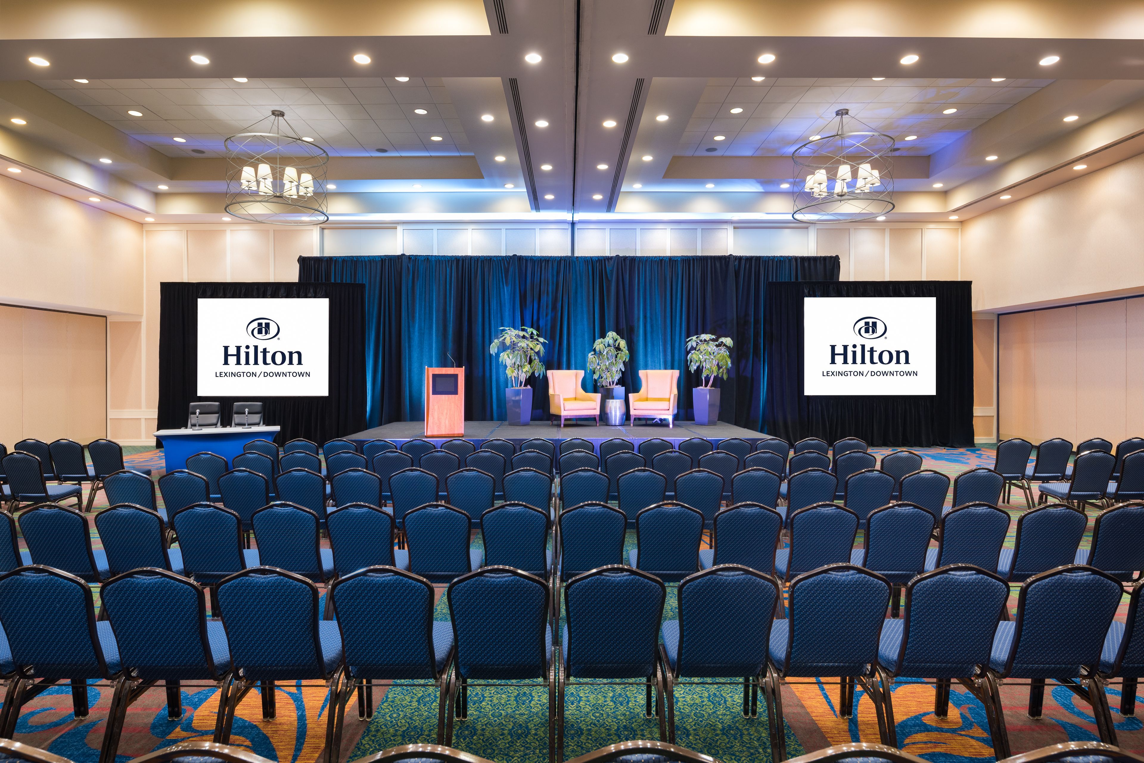 Een vergaderzaal met rijen blauwe stoelen, een podium en twee grote schermen waarop het Hilton-logo staat.