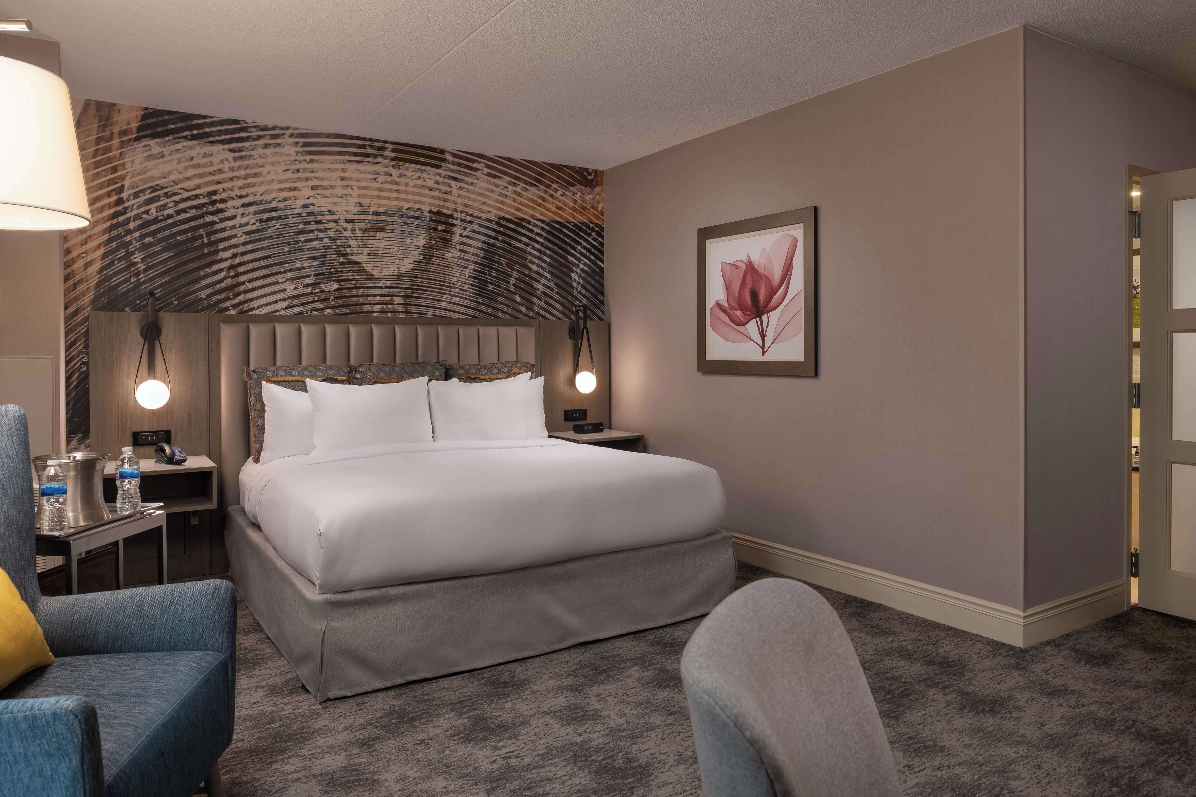 Habitación de hotel con una cama grande, dos mesitas de noche, una silla, una lámpara y una obra de arte colgada en la pared.