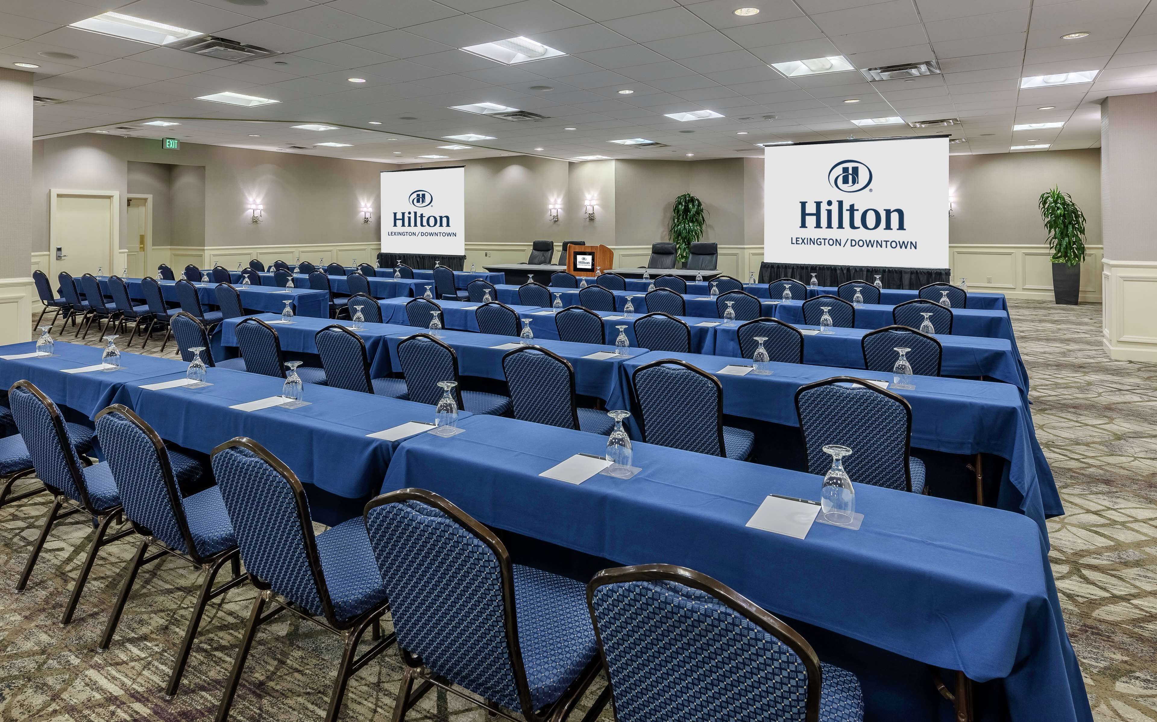 A conference room with rows of tables and chairs, a podium, and a projector screen.