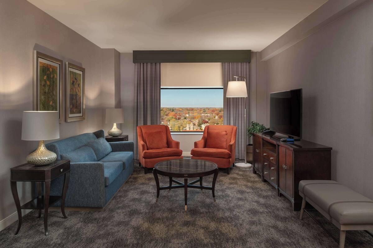 Quarto de hotel moderno com sofá azul, duas poltronas vermelhas, mesa de centro redonda, televisão e vista para a paisagem urbana através de grandes janelas.