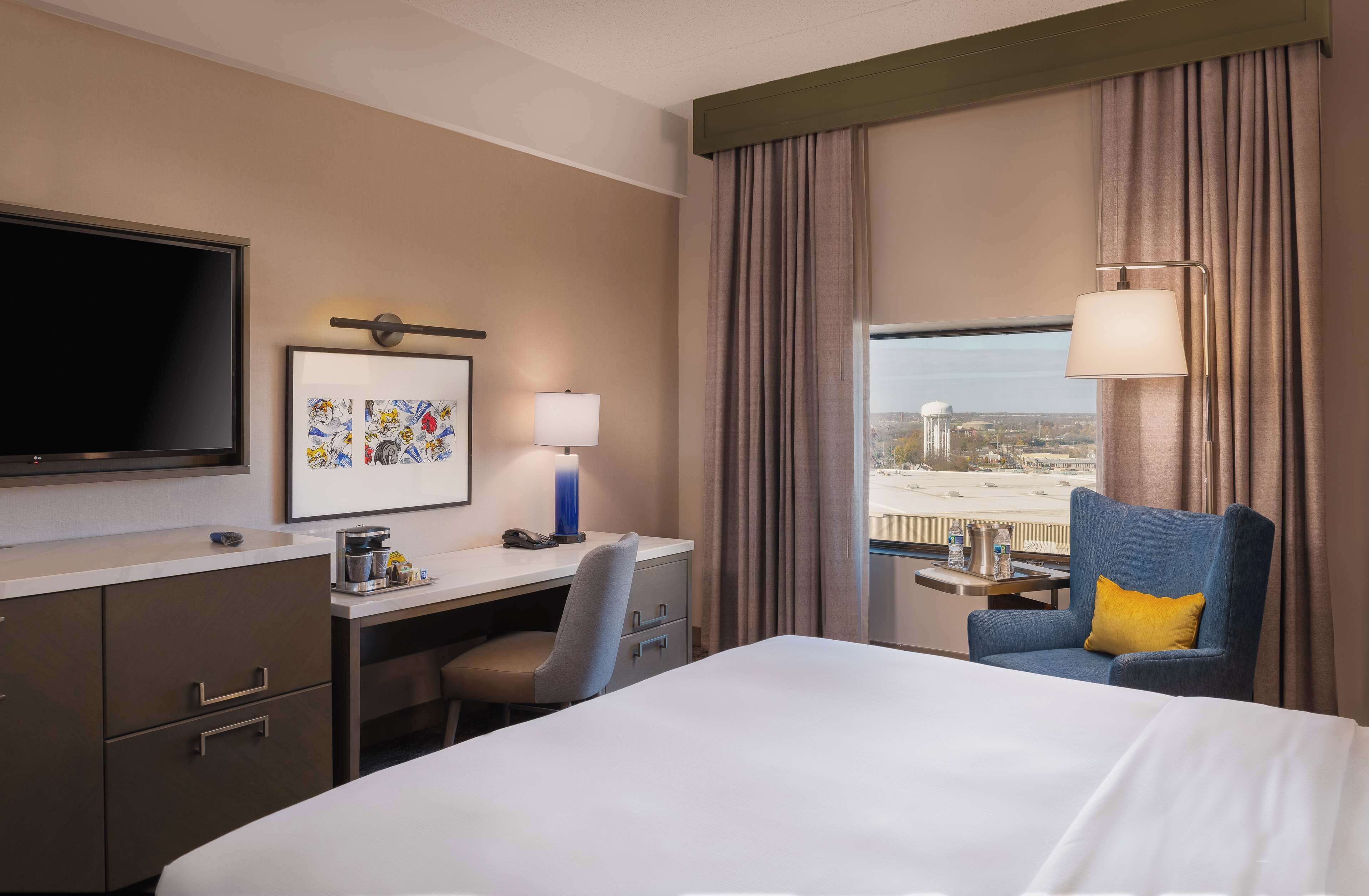 Ein Hotelzimmer mit Bett, Schreibtisch, Stuhl, Fernseher und einem Fenster mit Vorhängen.