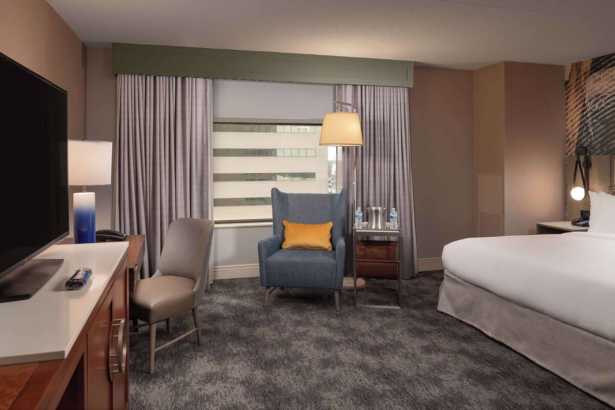 Quarto de hotel com cama, escrivaninha com televisão, cadeira, abajur e janela com cortinas.