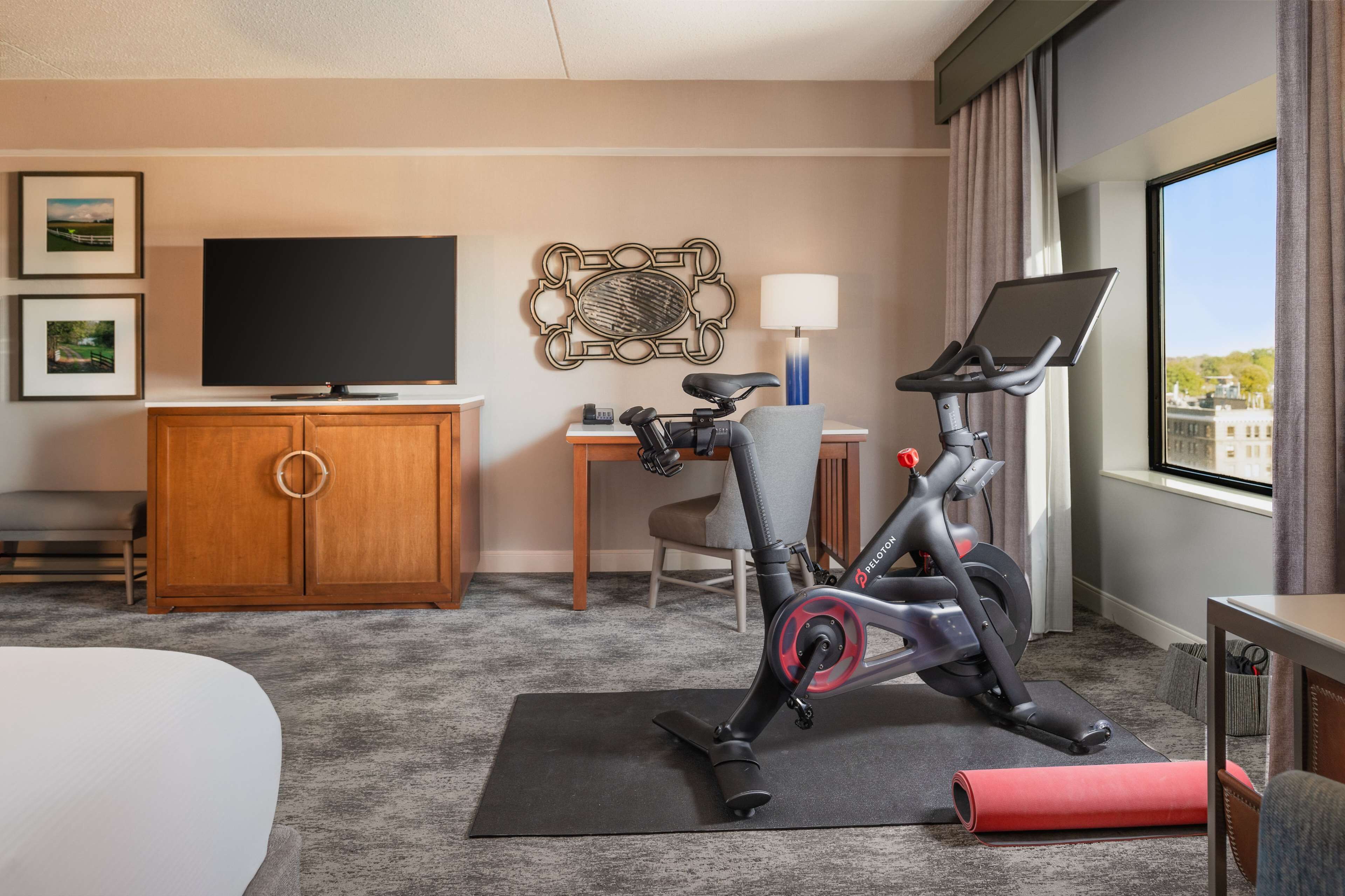 Ein Hotelzimmer mit Bett, Ergobike, Schreibtisch, Stuhl, Fernseher und Blick auf Gebäude durch das Fenster.
