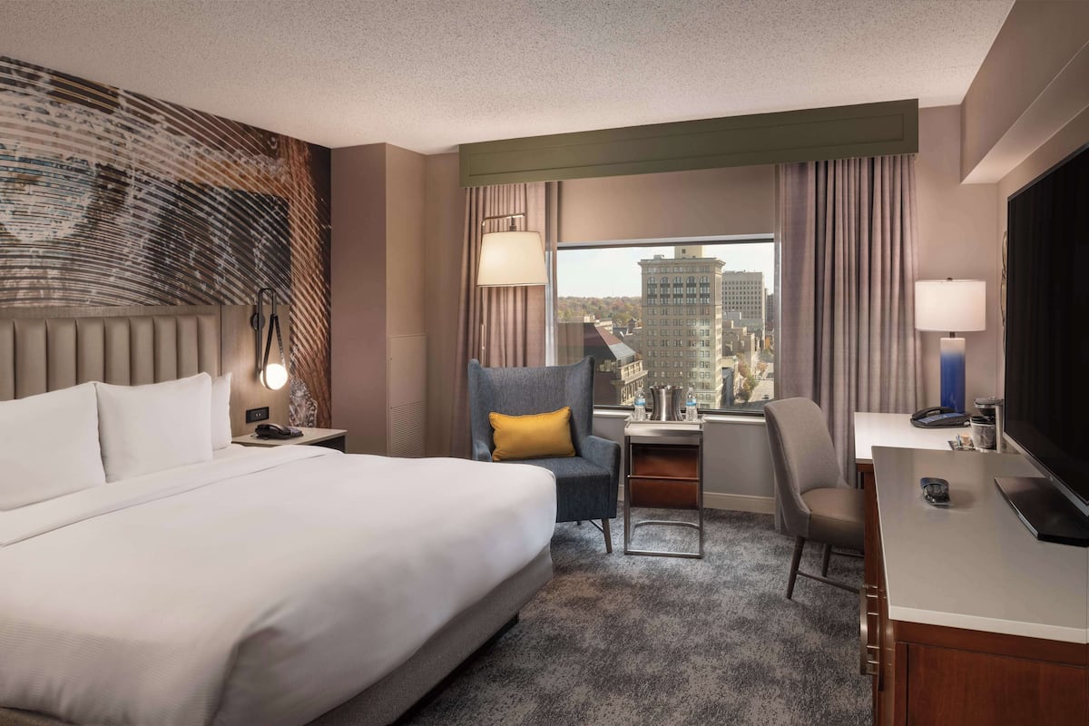 Quarto de hotel com uma cama grande, uma escrivaninha com cadeira, televisão e vista para a cidade pela janela.