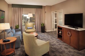 A hotel room with a sofa, armchair, small table, TV, cabinet, and a view of the city through the window.