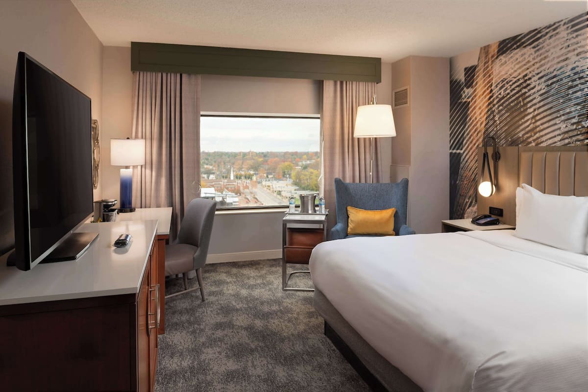 Quarto de hotel com cama, escrivaninha, cadeira, televisão e vista para a cidade pela janela.