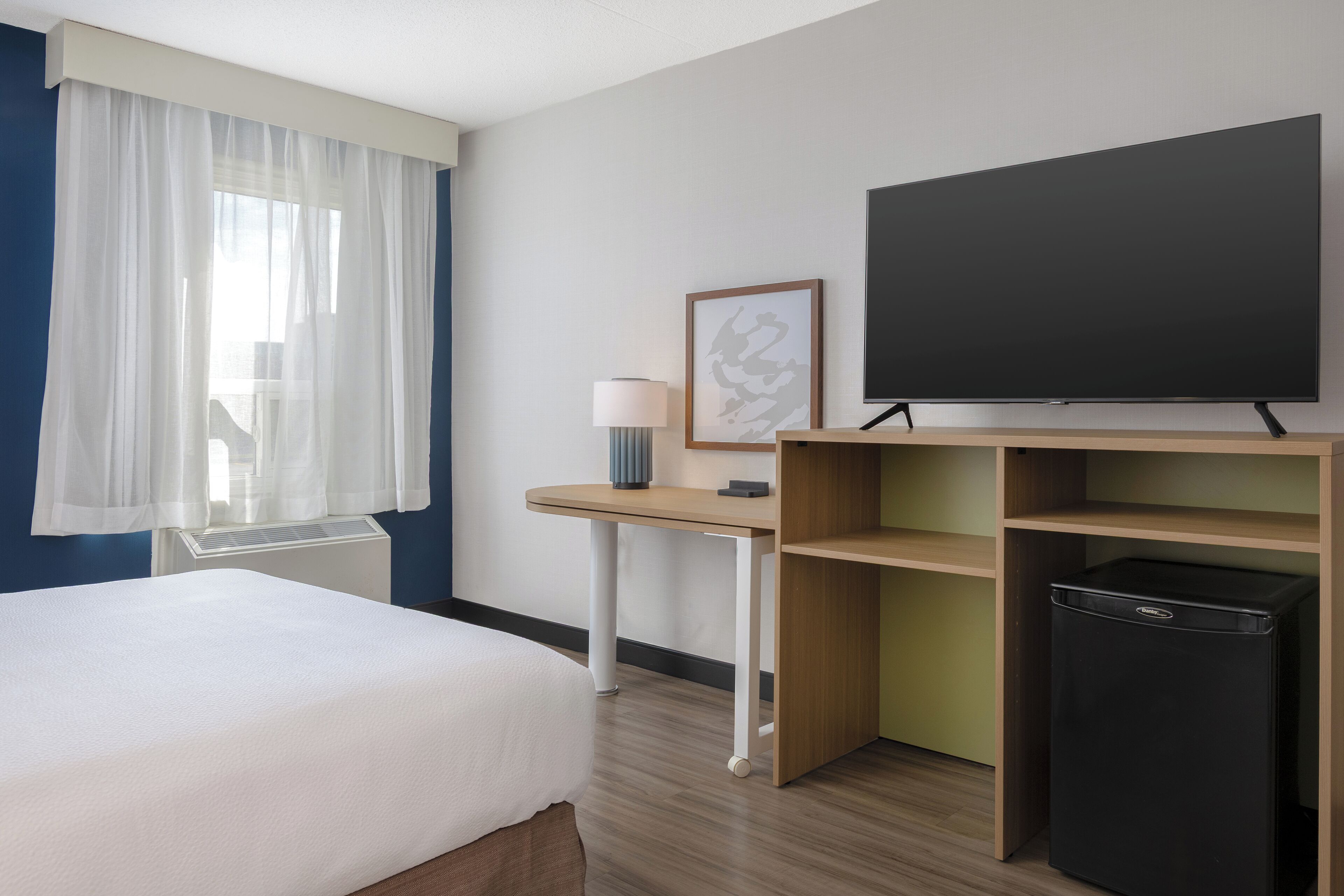 A hotel room with a bed, a desk, a television, a lamp, and a small refrigerator.