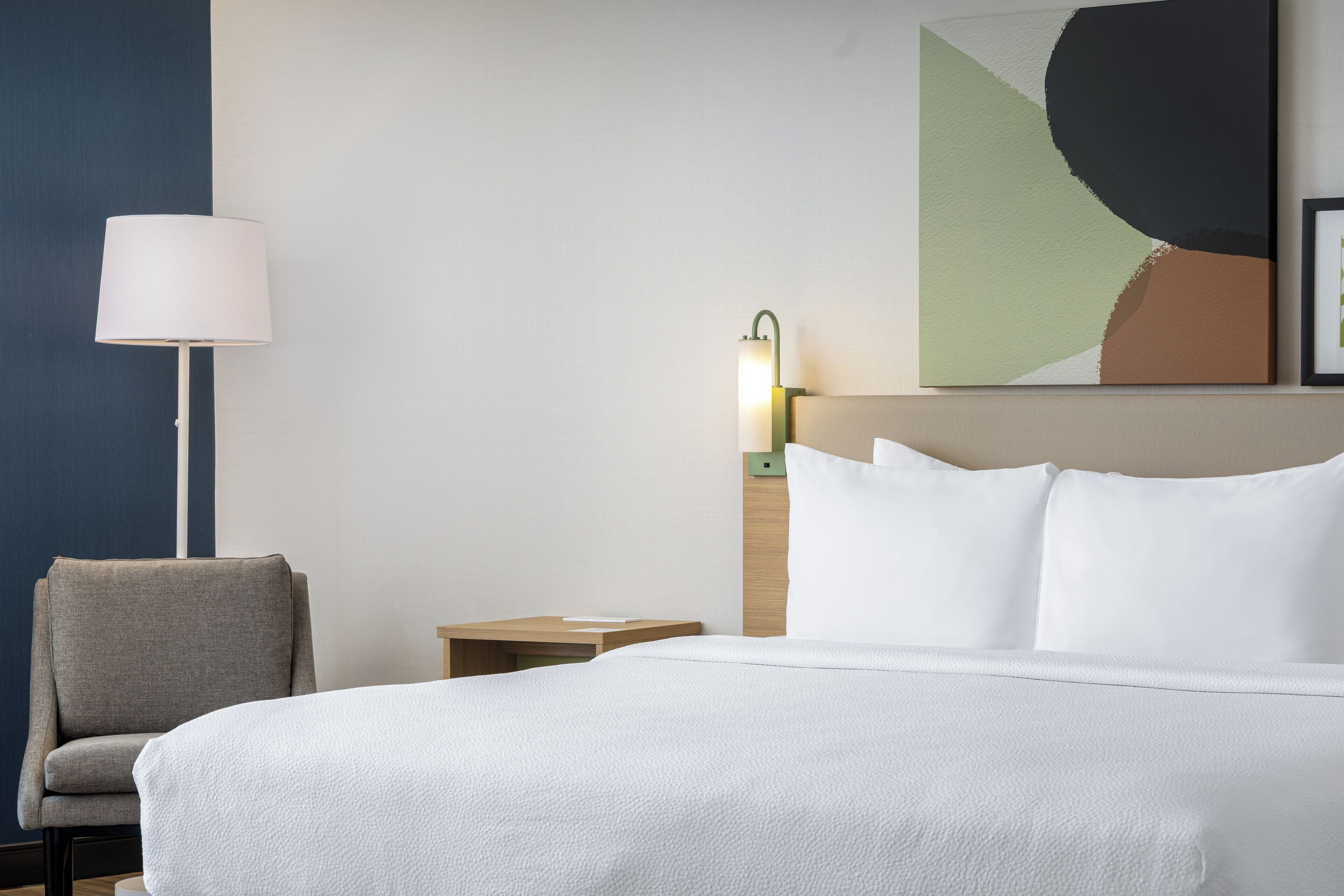 Una habitación de hotel con una cama, una silla, una mesita de noche, una lámpara y una obra de arte en la pared.