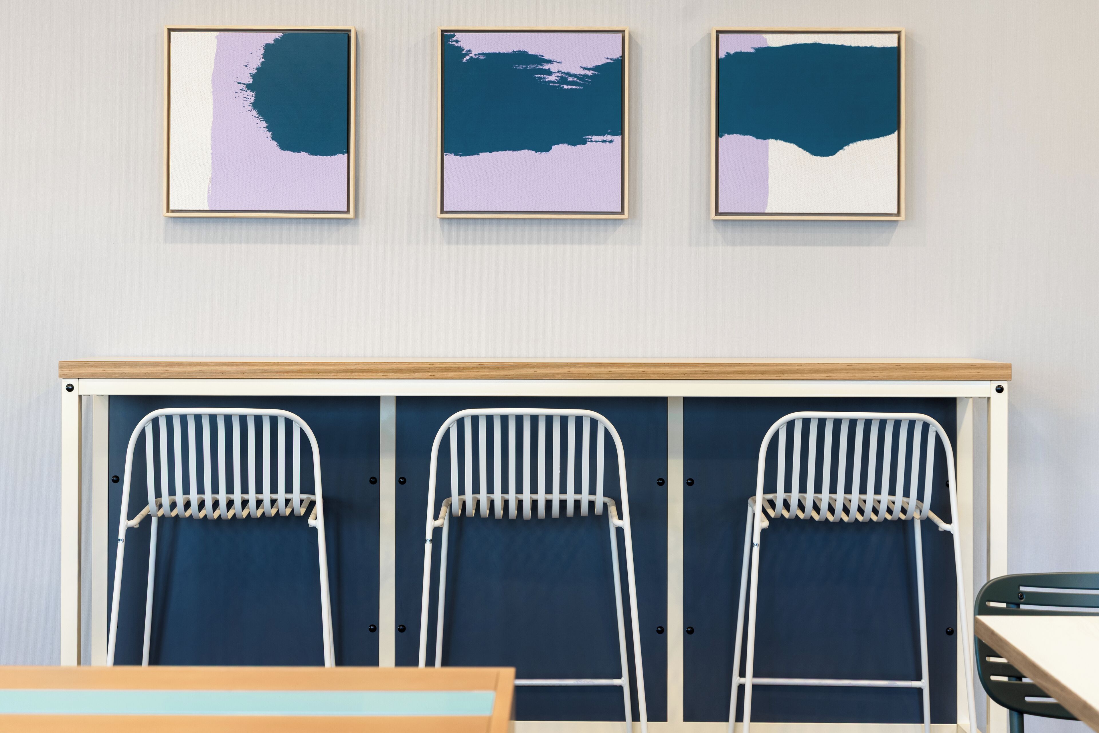 A room with three framed abstract paintings, a bunk bed with two white metal chairs, and a wooden table.
