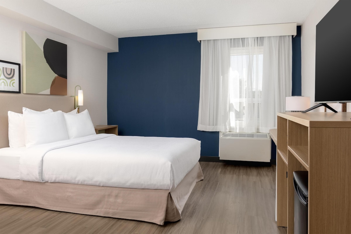 Ein Hotelzimmer mit Bett, Schreibtisch, Stuhl, einem Fenster mit Vorhängen, einem an der Wand montierten Fernseher und einer Nachttischlampe.