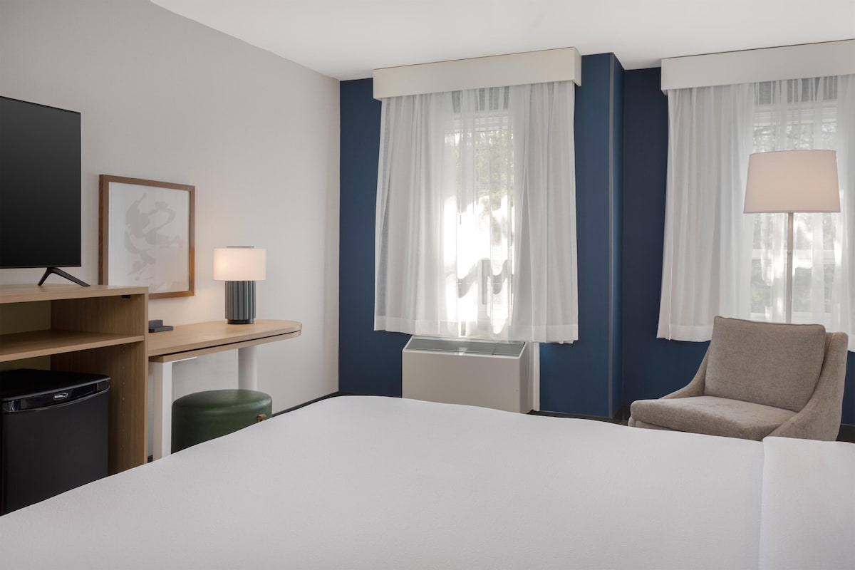 Ein Hotelzimmer mit einem Bett, einem Sessel, einem Schreibtisch mit Lampe, einem Fernseher und einem Bild an der Wand.