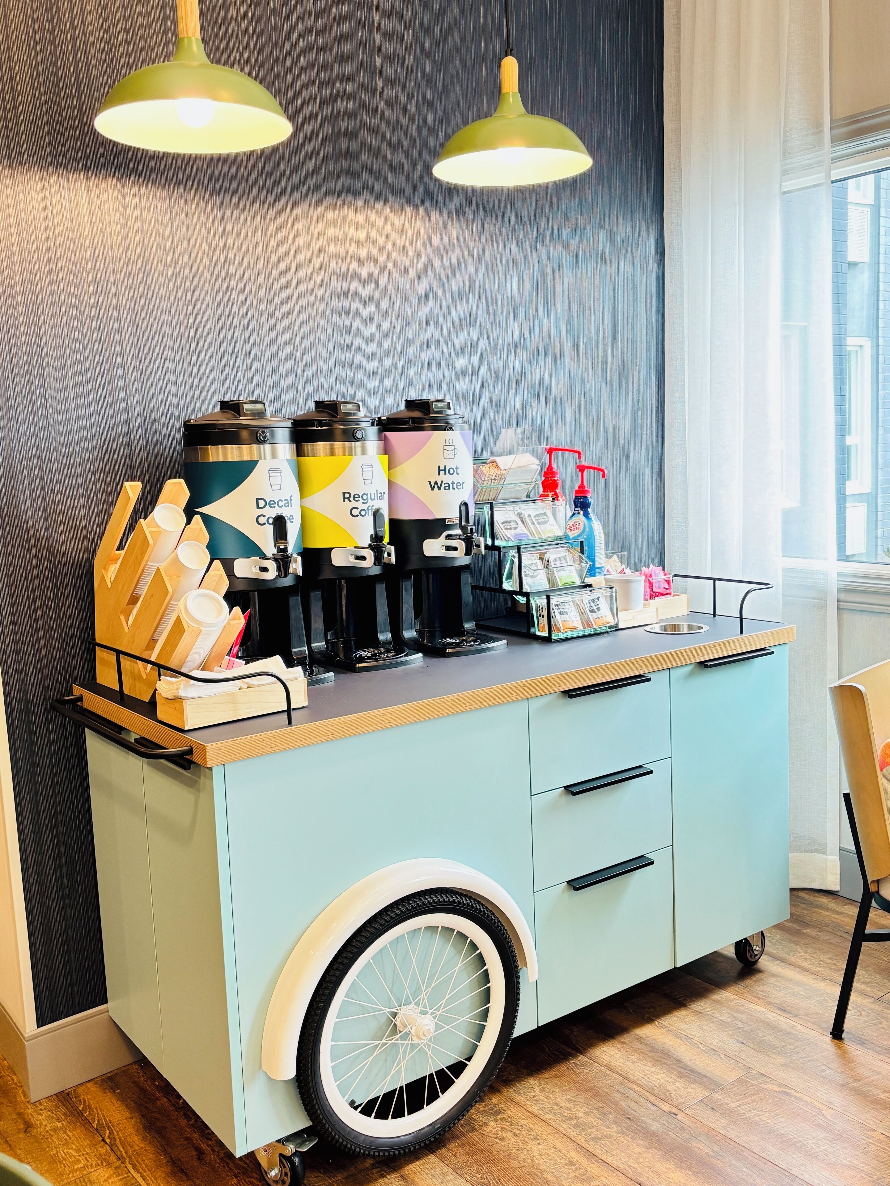 A coffee station with a vintage cart, two coffee dispensers, a hot water dispenser, and a small selection of condiments and napkins.