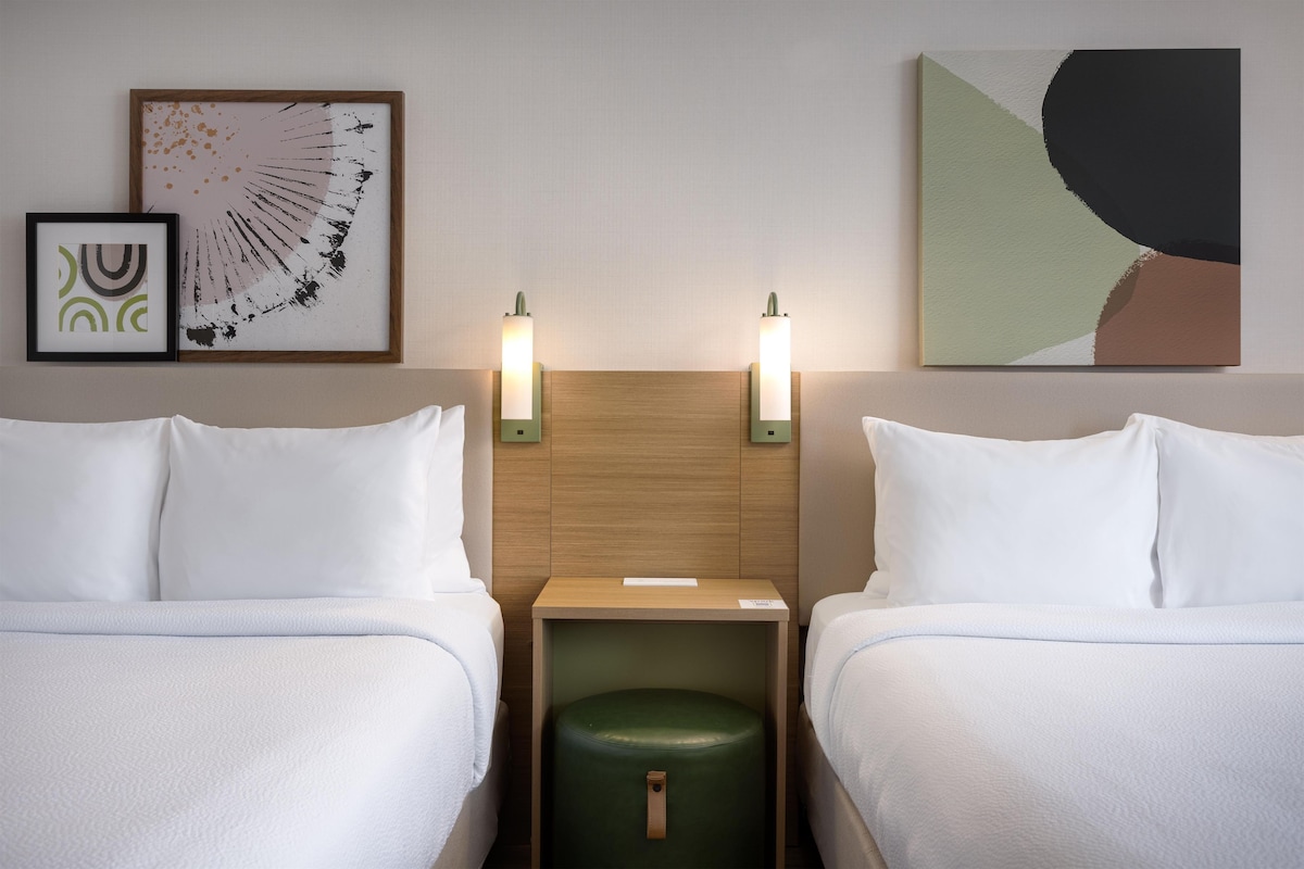 A hotel room with two beds, a wooden headboard, bedside lamps, and artwork on the walls.