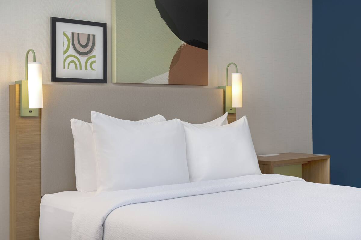 A neatly made bed with white linens and pillows, two wall-mounted lamps, and a framed abstract artwork above the bed.