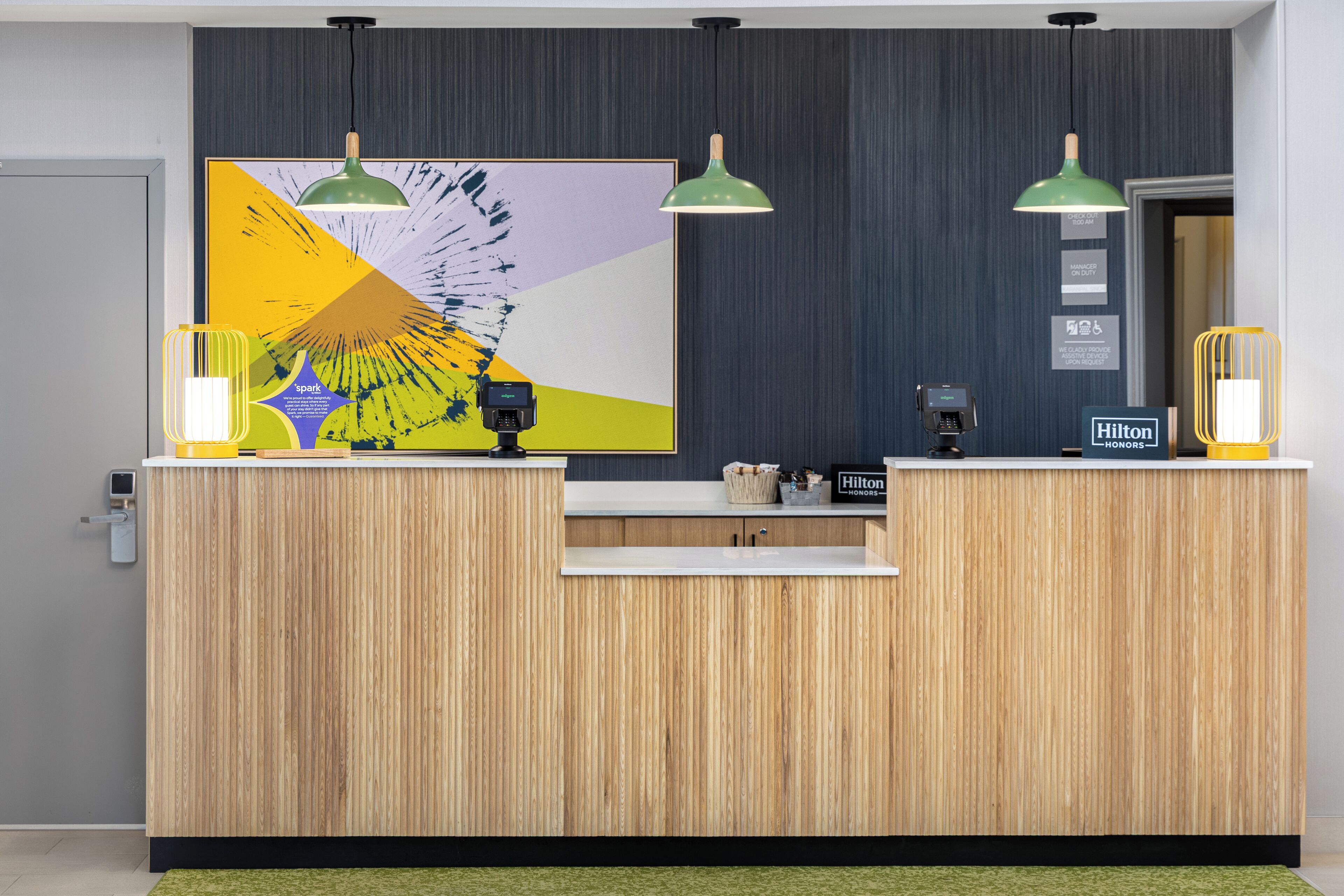 A wooden reception desk with a large abstract painting, two green pendant lights, a minibar, and a sign indicating Hilton Honors membership.