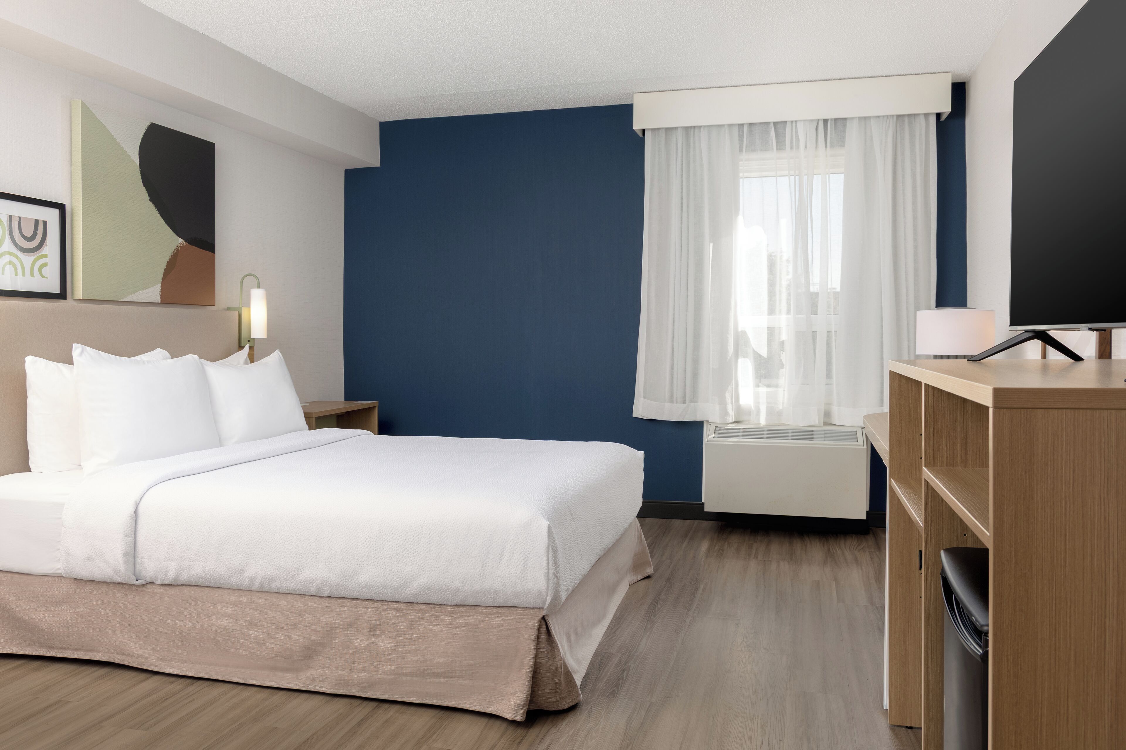 A hotel room with a bed, a desk, a chair, a lamp, a window with curtains, and a wall with a painting.