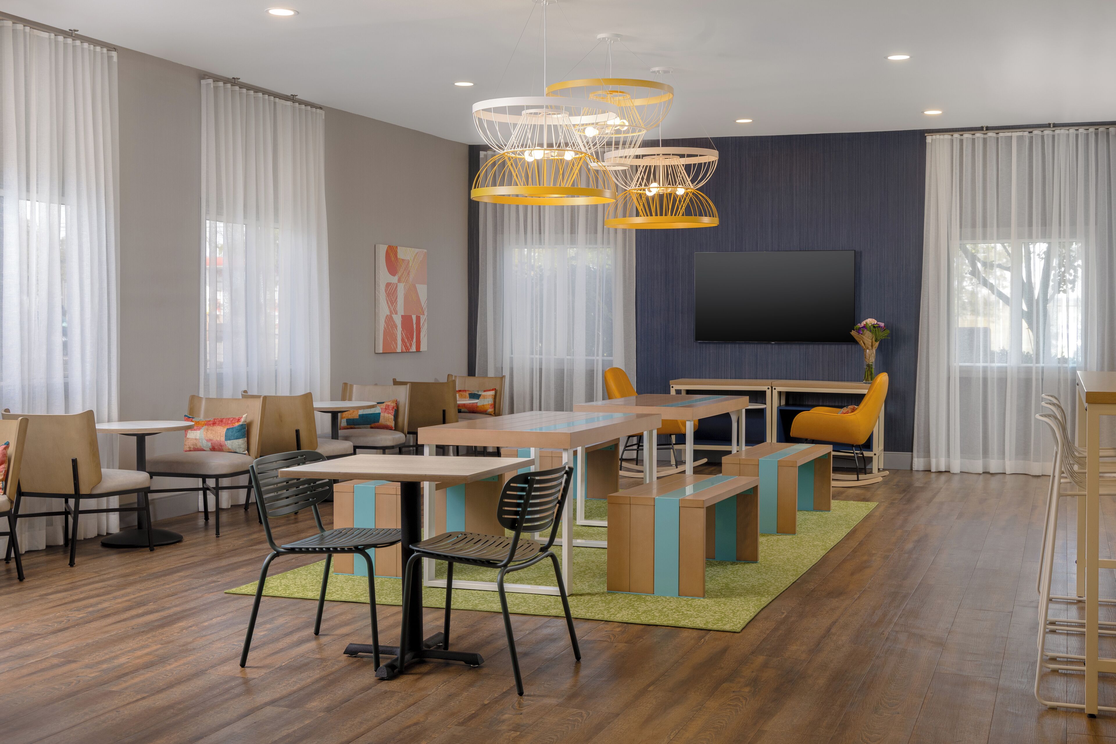 A modern dining area with wooden tables and chairs, a flat-screen TV, and a large window with sheer curtains.