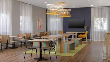 A modern dining area with wooden tables and chairs, a flat-screen TV, and a large window with sheer curtains.
