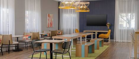 A modern dining area with wooden tables and chairs, a flat-screen TV, and a large window with sheer curtains.