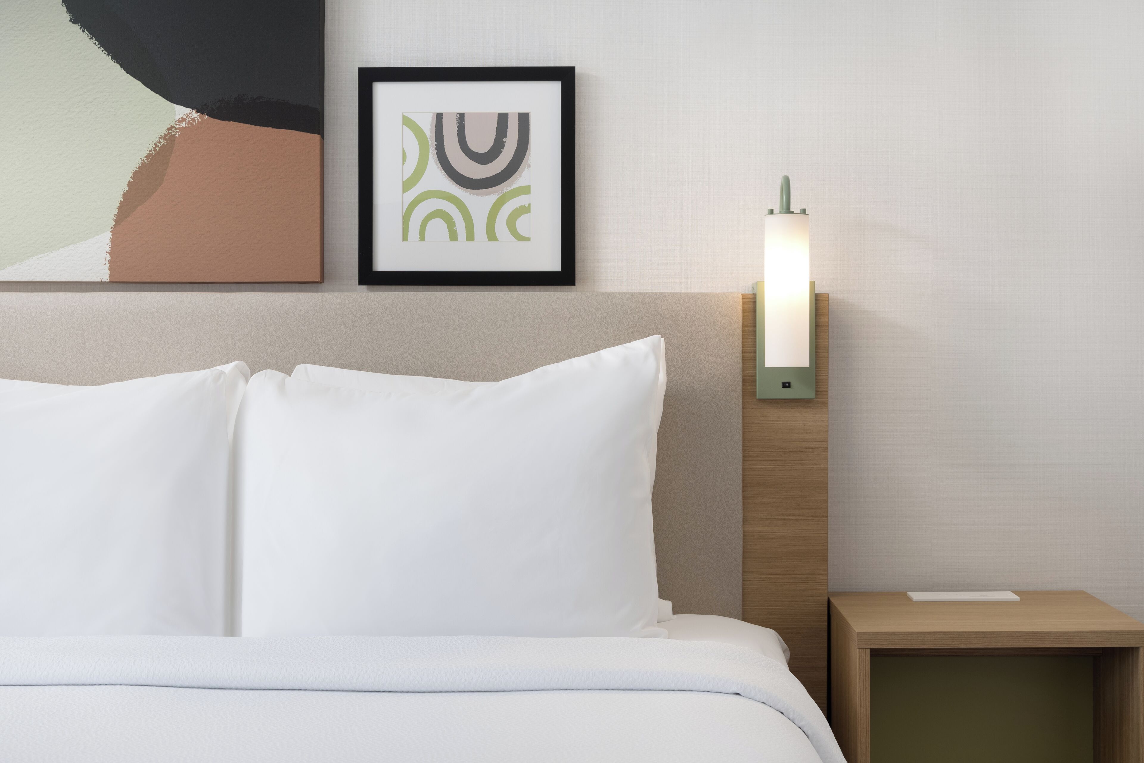 A neatly made bed with white linens, a framed abstract artwork above it, and a bedside table with a lamp.