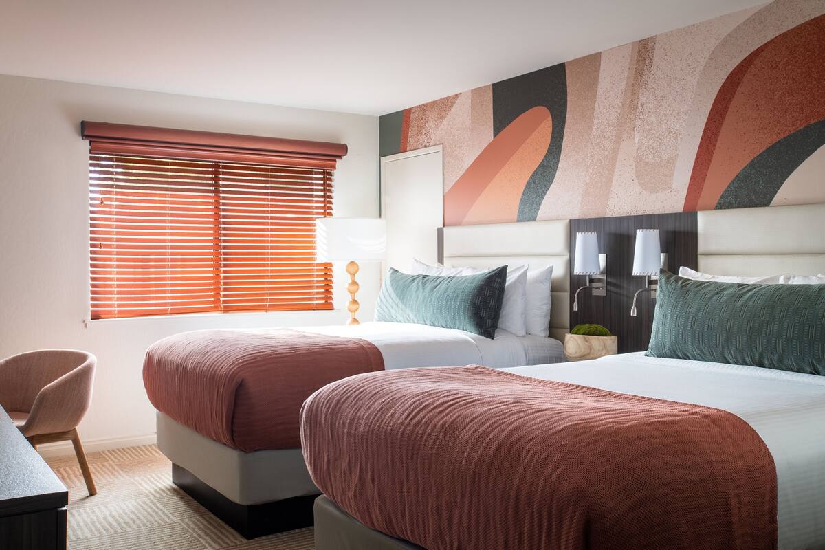 A hotel room with two beds, a desk, and a window with blinds.