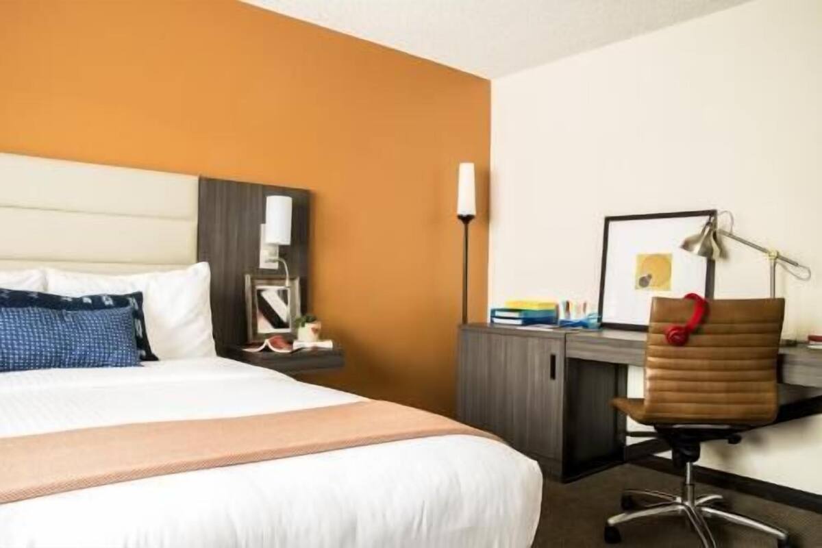 A hotel room with a bed, a desk, a chair, a lamp, and a framed picture on the wall.