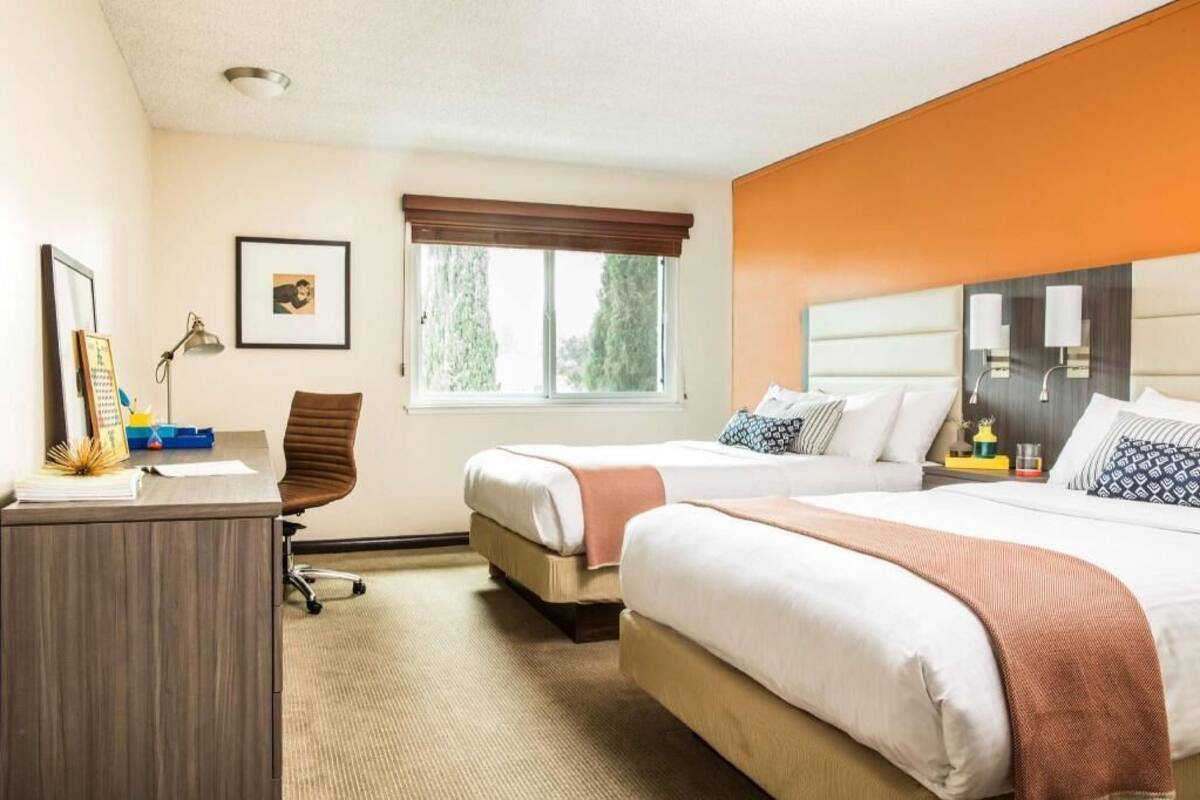 A hotel room with two beds, a desk, a chair, a window with a view, and a headboard with lamps.