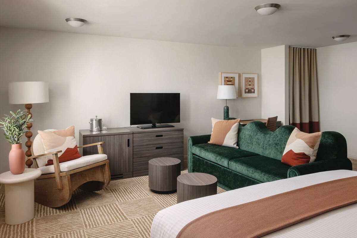 A modern hotel room with a green sofa, a wooden chair, a flat-screen TV, a small side table with a vase, and a patterned rug.