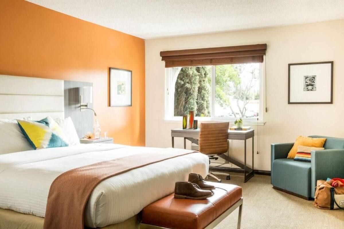 A hotel room with a bed, a desk, a chair, a window with a view of trees, and a sofa.