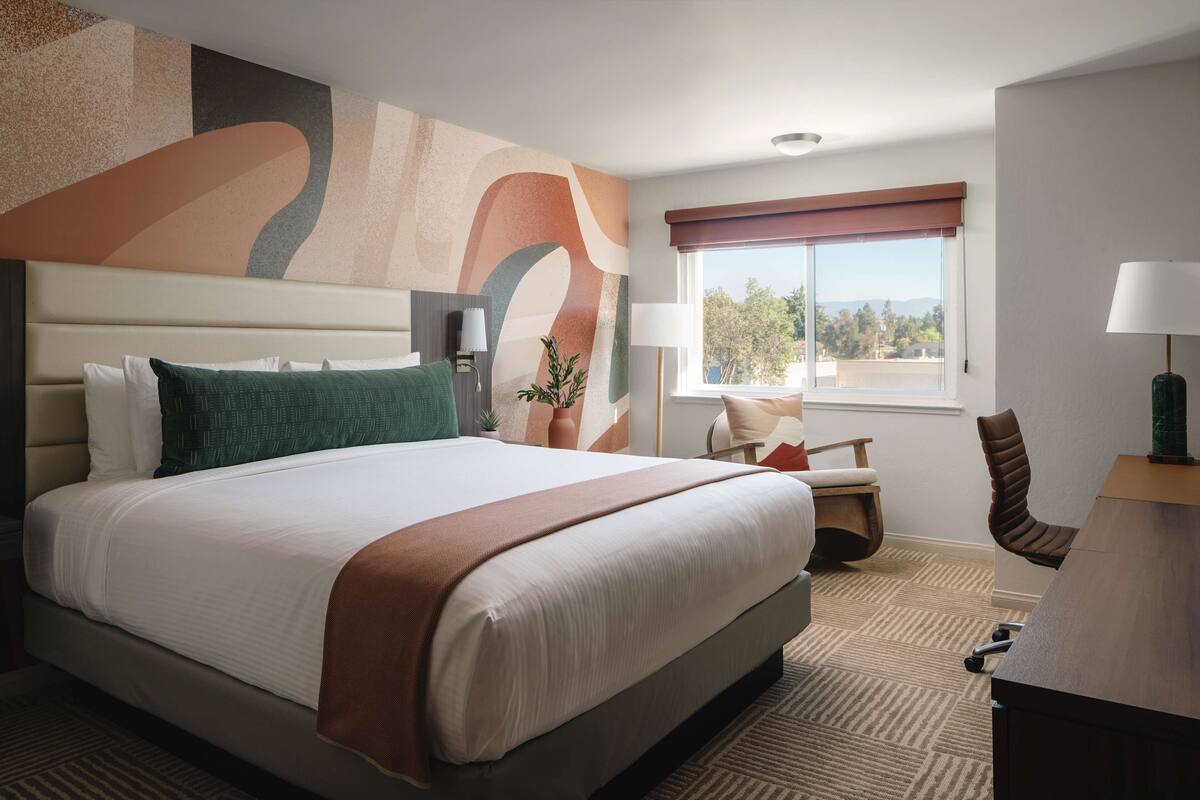 A hotel room with a large bed, a desk, a chair, a window with a view, and a wall with abstract art.