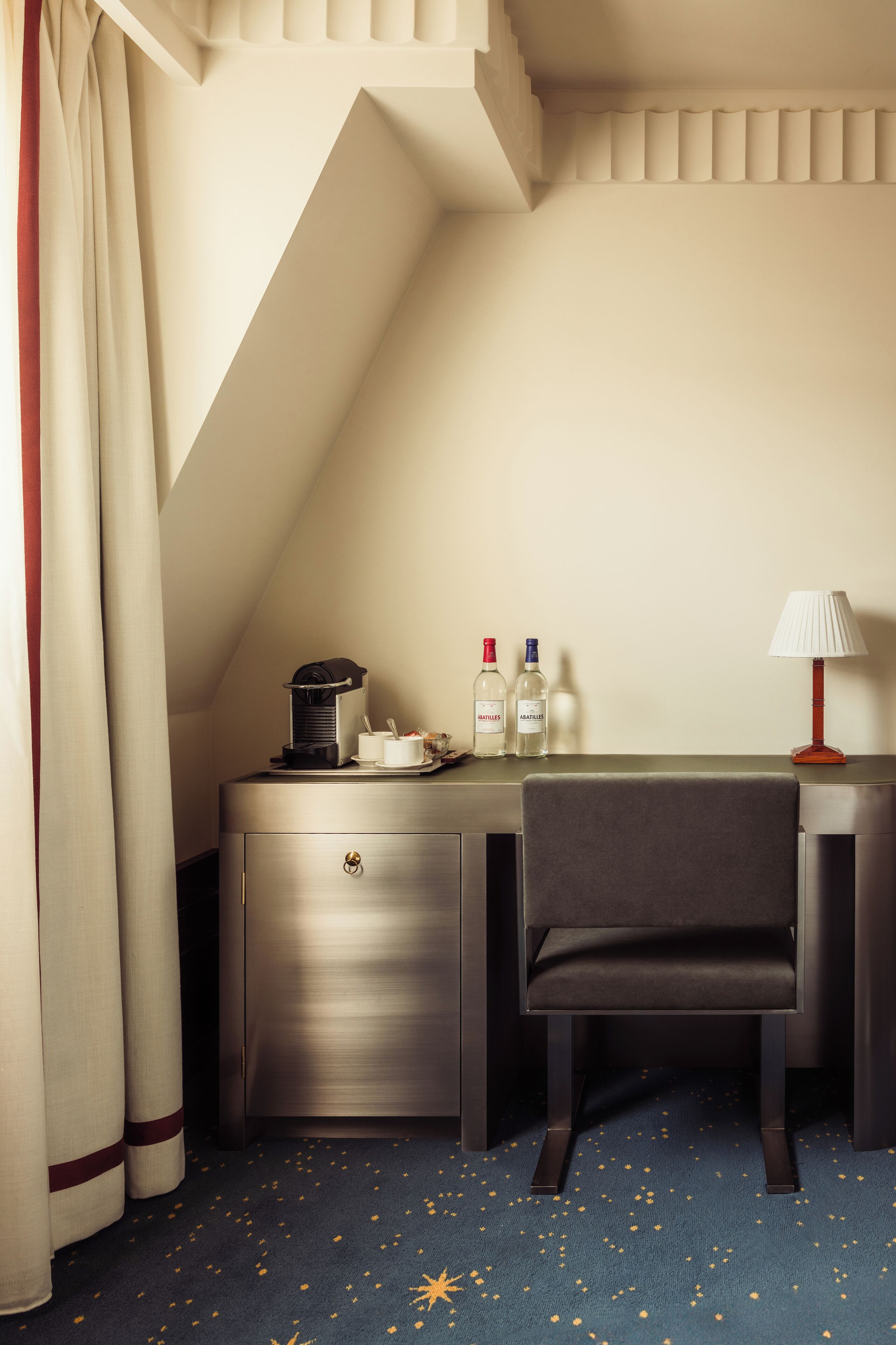 A hotel room with a desk, chair, and a small table with a coffee maker, two bottles, and a lamp.