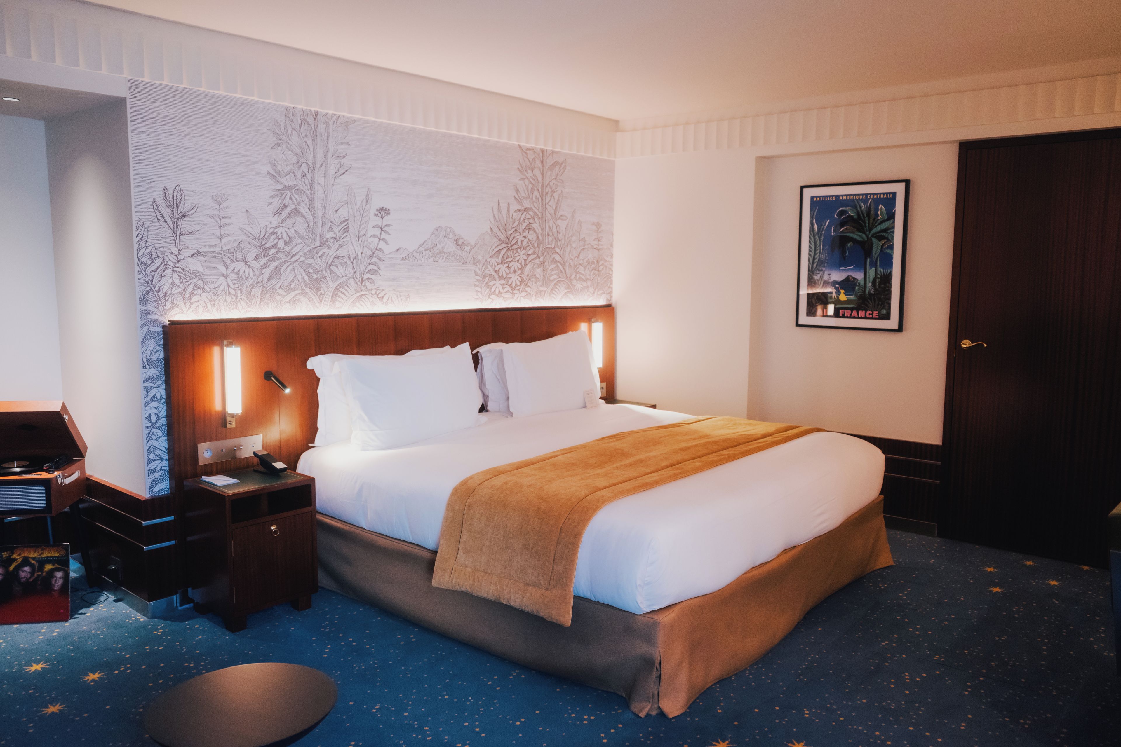 A hotel room with a large bed, a nightstand, a desk with a computer, and a wall with a poster.