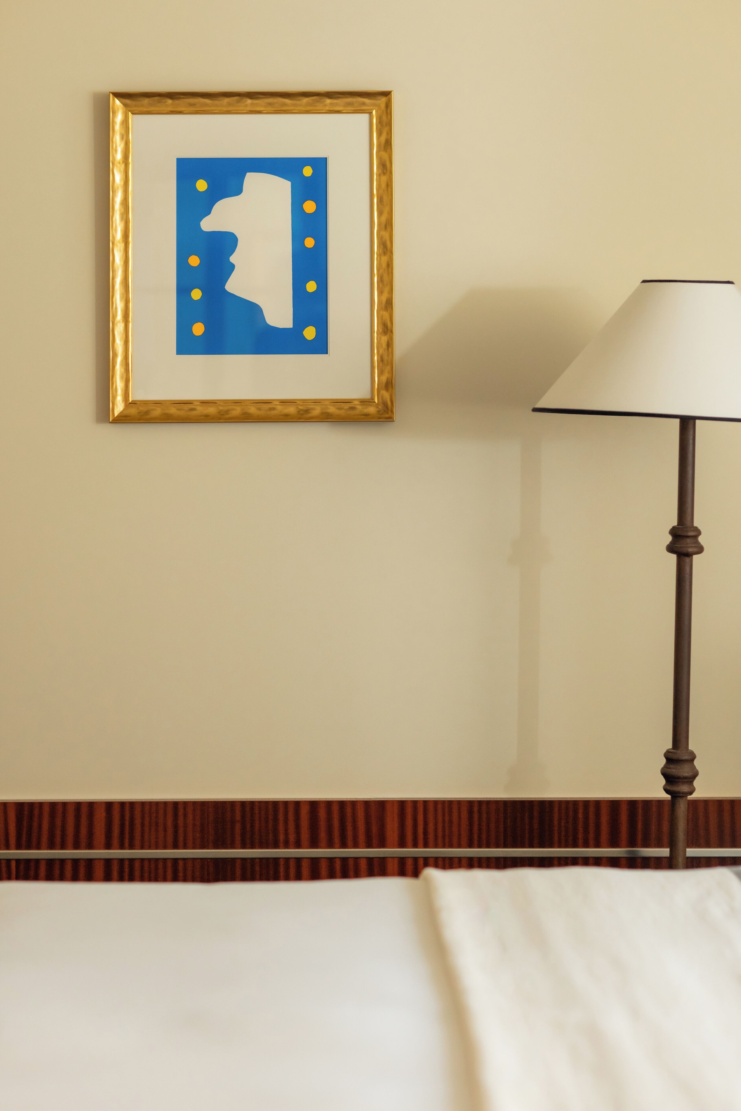 Una obra de arte enmarcada en una pared, una lámpara y una cama con cubrecolor blanco.