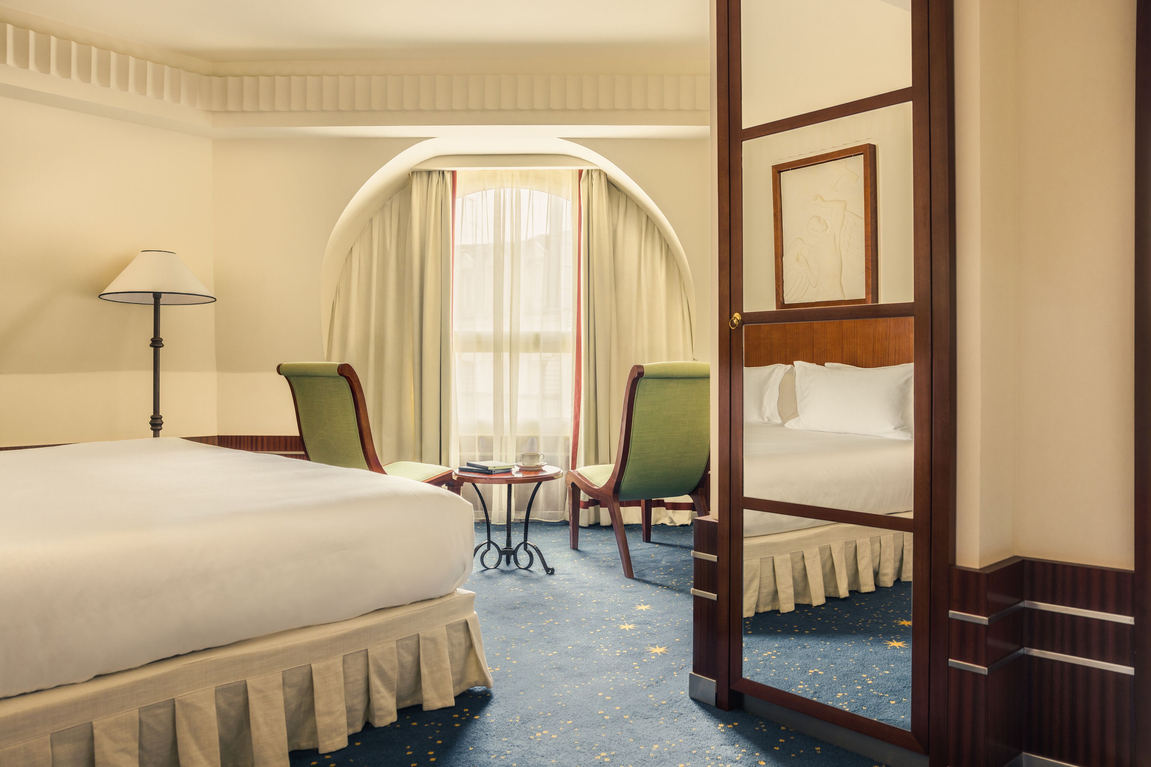 A hotel room with a large bed, two chairs, a small table, and a window with curtains.
