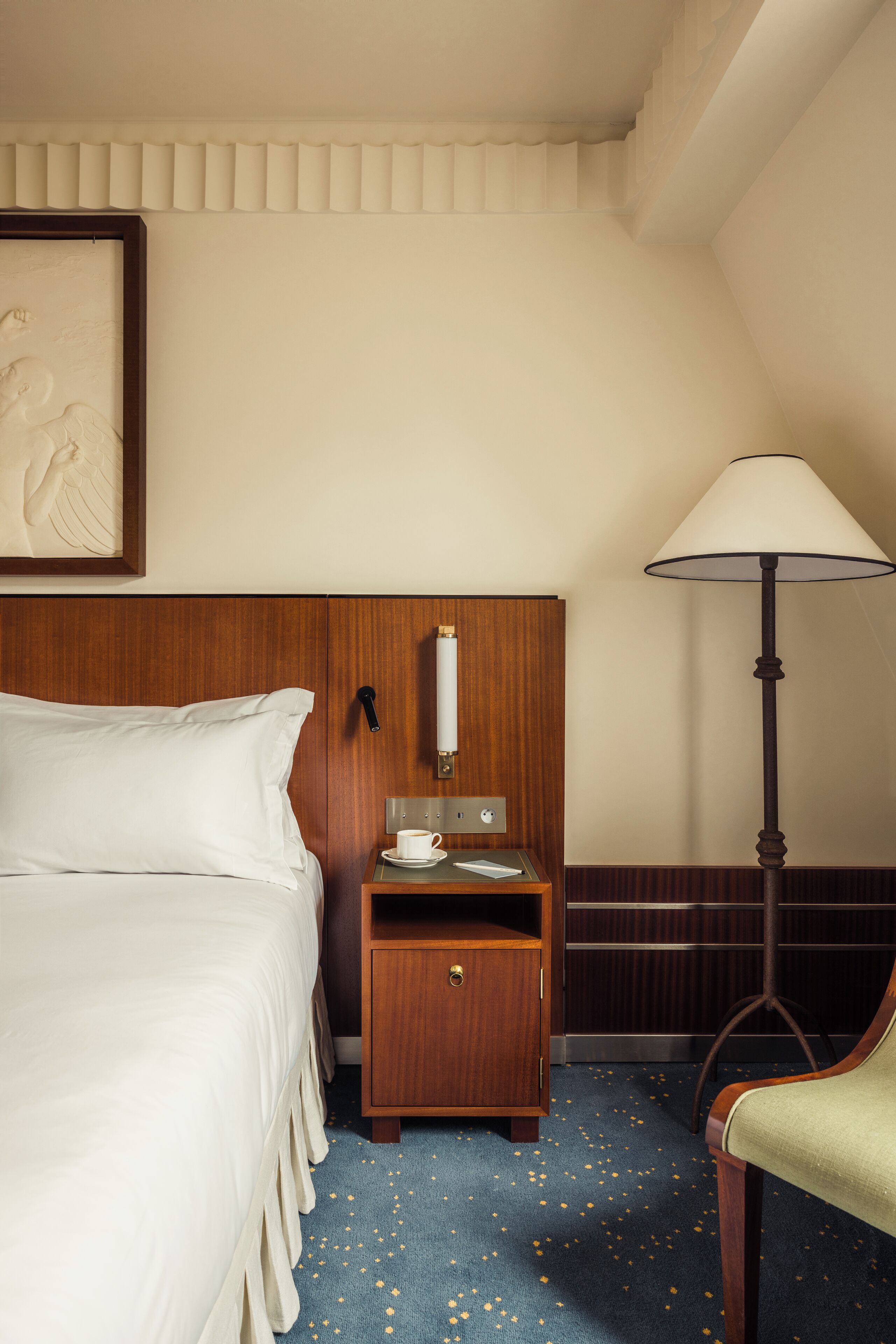 Une chambre d’hôtel avec un lit bien fait, une table de chevet en bois sur laquelle se trouvent une tasse et une lampe, et une chaise.