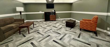 A living room with a herringbone carpet, a brown sofa, a wooden coffee table, an orange armchair, a TV mounted on the wall, and a small cabinet with drawers.